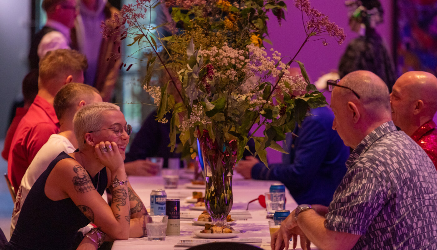 People sit around a long, rectangular table engaged in conversation at a social event. The table is decorated with a large, colorful floral arrangement in a tall vase. The atmosphere is lively, with several attendees smiling and chatting. The setting is warmly lit with a pinkish hue, adding to the vibrant ambiance of the gathering. Drinks and small plates of food are visible on the table. A woman with short blonde hair, tattoos, and glasses is prominently featured in the foreground, leaning forward and smiling.