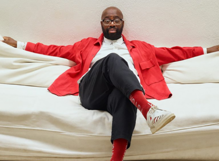 A black, bald man with a beard and glasses is sitting on a white sofa, dressed in a bright red jacket, white shirt, black pants, and red socks with white sneakers. He is lounging with his arms stretched out along the back of the sofa, exuding a relaxed and confident demeanor. The background is a plain, light-colored wall.