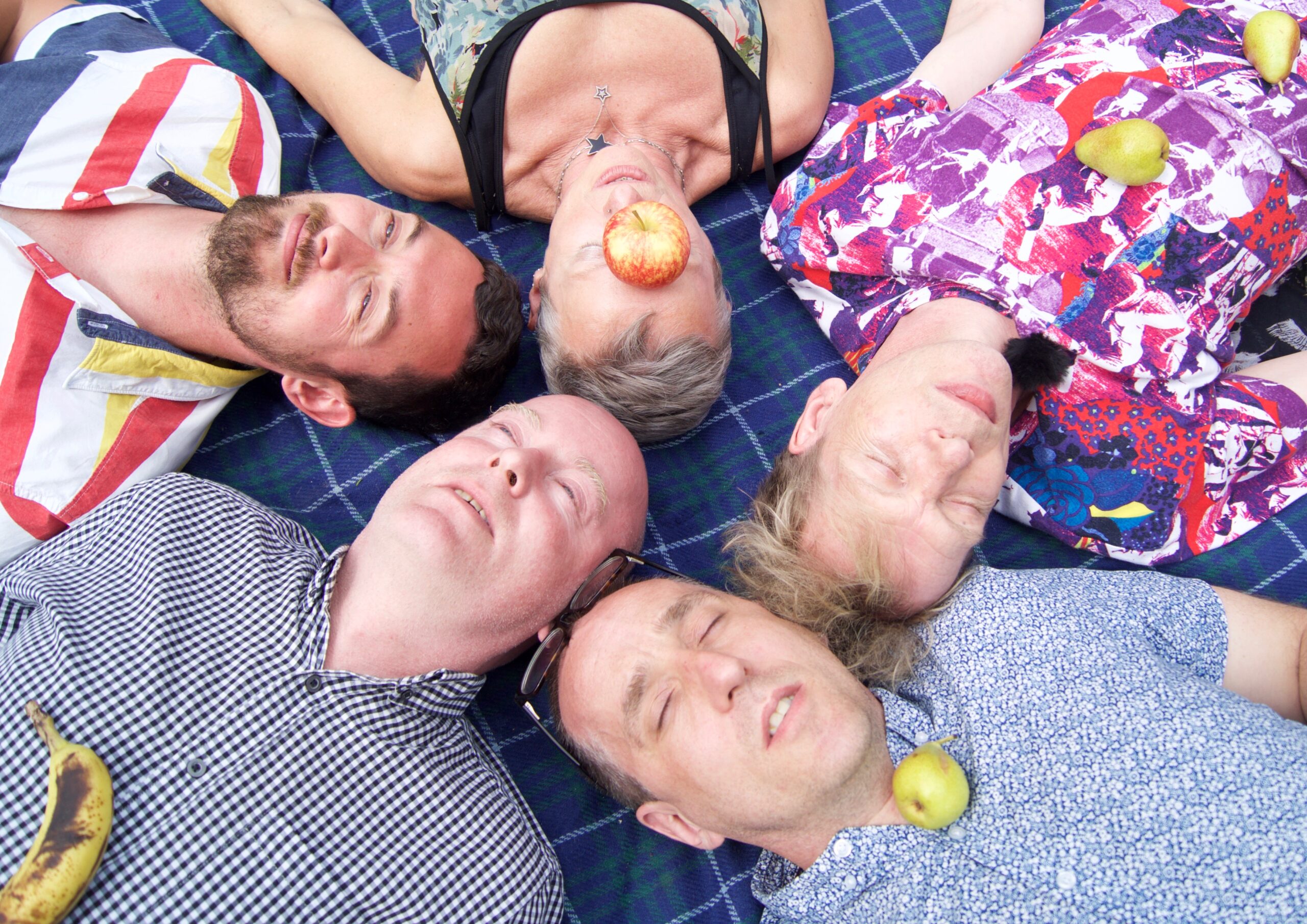 Five people lie on their backs in a circular arrangement on a blue plaid blanket. They appear relaxed, with eyes closed. The person in the center has a red apple placed over their mouth, while the others have fruits such as a banana and pears resting on their clothing. Their outfits are colorful and varied, adding to the playful and relaxed atmosphere of the scene.